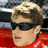 A static image shows a person with short light brown hair wearing dark sunglasses and a red shirt against a red background with white text.