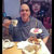 A pixelated static image shows a person in a dark blue shirt holding a white plate with food, seated at a table with a light-colored surface.