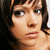 A pixelated close-up of a person with brown hair and light skin, showing eyes, nose, mouth, and part of the shoulder against a dark background.