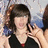 Pixelated image of a person with dark hair tied in a high ponytail, wearing a black top and red necklace against a blurred background.
