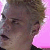 Pixelated close-up of a person's face with light brown hair against a pink background, showing partial facial features and clothing.