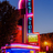 Pixelated image of a tall building with vertical color bands against a blue sky, dark tree silhouette on left, and warm-toned ground elements.