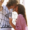 Two people kiss near a tennis net; one wears a white shirt with black lines, the other a pink patterned shirt.