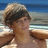 Pixelated image of a person with dark hair wearing a light-colored hat, standing against a beach background with blue sky and water.