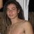 A shirtless person with long brown hair stands against a dark background with white vertical elements, expression neutral.