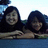 Two people with dark hair lean forward on a blue surface, smiling at the camera against an outdoor background.