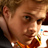 Pixelated close-up of a person's face with light brown hair, wearing an orange and white patterned garment against a dark background.