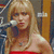 A pixelated static image shows a person with long blonde hair wearing a red strapless top, holding a microphone near their mouth against a blurred background.