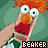 Pixelated character with orange hair, large white eyes, and red mouth holding a green object against teal background.