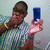 A person in a striped shirt holds a blue phone, wearing a yellow bracelet and a green-and-purple wristband, with a light-colored wall background.