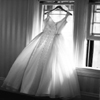 A white wedding dress hangs on a hanger in front of a window with vertical blinds, illuminated by bright light from the window.