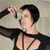Pixelated image of a person with short dark hair wearing a black strappy top, raising one arm with a hand gesture against a light background.