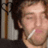 A pixelated close-up of a person with light skin holding a white cigarette near their mouth, set against an indistinct background.