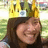 A person with long dark hair wears a golden crown against a blurred green and yellow background.