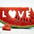 A pixelated red watermelon slice with green rind and black seeds, featuring the word LOVE where O is a white heart shape.