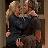 Two figures in close embrace, one with light hair, the other with darker hair, both wearing dark clothing against a blurred background.