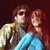 Two figures sit closely together; one wears sunglasses and a light jacket, the other has long red hair and blue pants, with partial text at the bottom.