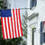 Static pixelated image of a red and white striped flag with a blue canton, positioned against a blurred background of green foliage and light-colored structures.