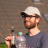 Pixelated image of a person wearing a white cap and dark shirt holding a bottle with a green cap against a blurred background.