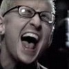 A close-up of a person with short light-colored hair wearing dark-framed glasses, mouth open in a shout or yell, teeth visible, ear visible, dark background.