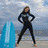 A woman in a black wetsuit stands on a beach with hands on hips, next to a blue surfboard under a cloudy sky.