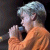 Pixelated image of a person with light blue hair holding a microphone, wearing an orange garment against a dark background.