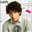 Pixelated image of a person with dark curly hair wearing a light gray jacket over a black shirt, standing against a white background with scattered pink and blue pixels.