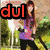 A woman with long brown hair in a purple shirt and black pants stands outdoors holding an object, with the text 'dul' displayed above her.