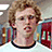 Pixelated image of a person with curly light brown hair wearing a white shirt with black collar, set against red background with repeated text.