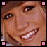 A pixelated close-up of a smiling person with light brown hair, wearing a dark cap and showing white teeth.