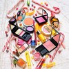 A static composition of various makeup products including eyeshadow palettes, lipsticks, and brushes arranged in a scattered pattern on a light background.