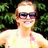 A person in a yellow top with pink accents stands outdoors against green foliage, wearing dark sunglasses and smiling, with a gray rectangular overlay on the right side.