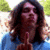 A person with long brown hair wearing a blue shirt holds up one finger in front of a blurred outdoor background.