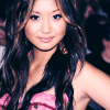 A woman with long wavy brown hair wears a pink strapless top with a small green detail, posing against a dark background.