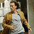 Pixelated image of a person wearing a white shirt with gray pattern and brown jacket, standing indoors near a doorway.