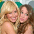 Two women with long hair pose closely together, one with blonde hair and the other with brown hair, both smiling against a blurred background.