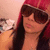 A person with long dark hair wears a red headscarf and large brown sunglasses, positioned against a light-colored wall with fabric items in the background.
