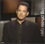Pixelated image of a person in dark clothing with a light-colored collar, seated against a blurred background with brown and gray tones.