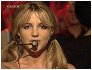 Britney Spears in a red dress with microphone, performing on stage with audience background.