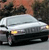 A silver sedan with yellow headlights is positioned on a road, viewed from a low angle with a blurred background of trees and sky.