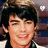 Pixelated close-up of a person with dark hair and a light-colored top, smiling with visible teeth against a blurred background.