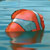 Pixelated static icon of a red-orange rounded object with smooth gradients against a blue striped background, appearing to be a stylized cartoon character or mascot head.