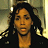 Pixelated static image of a person with long dark hair wearing a black jacket and large hoop earrings against a blurred background.