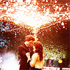 A couple embraces under a bright light source with a blurred background of trees and lights.