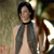 A pixelated figure with dark hair wears a sleeveless top and a choker, standing against a blurred background with green and brown tones.