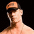 A shirtless man wearing an orange and black cap with a small dark rectangle on the front, standing against a solid black background.