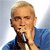 Pixelated image of a person with short light brown hair wearing a blue shirt, holding a microphone near their mouth against a light blue background.