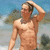 A shirtless man with short brown hair stands outdoors, hand raised to his head, against a blurred background of blue sky and green foliage.