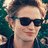 Pixelated close-up of a person with light brown hair wearing dark sunglasses and a dark blue jacket against a green background.