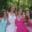 Three people stand closely together outdoors under green foliage, wearing light blue, white, and pink dresses with visible arm and shoulder details.